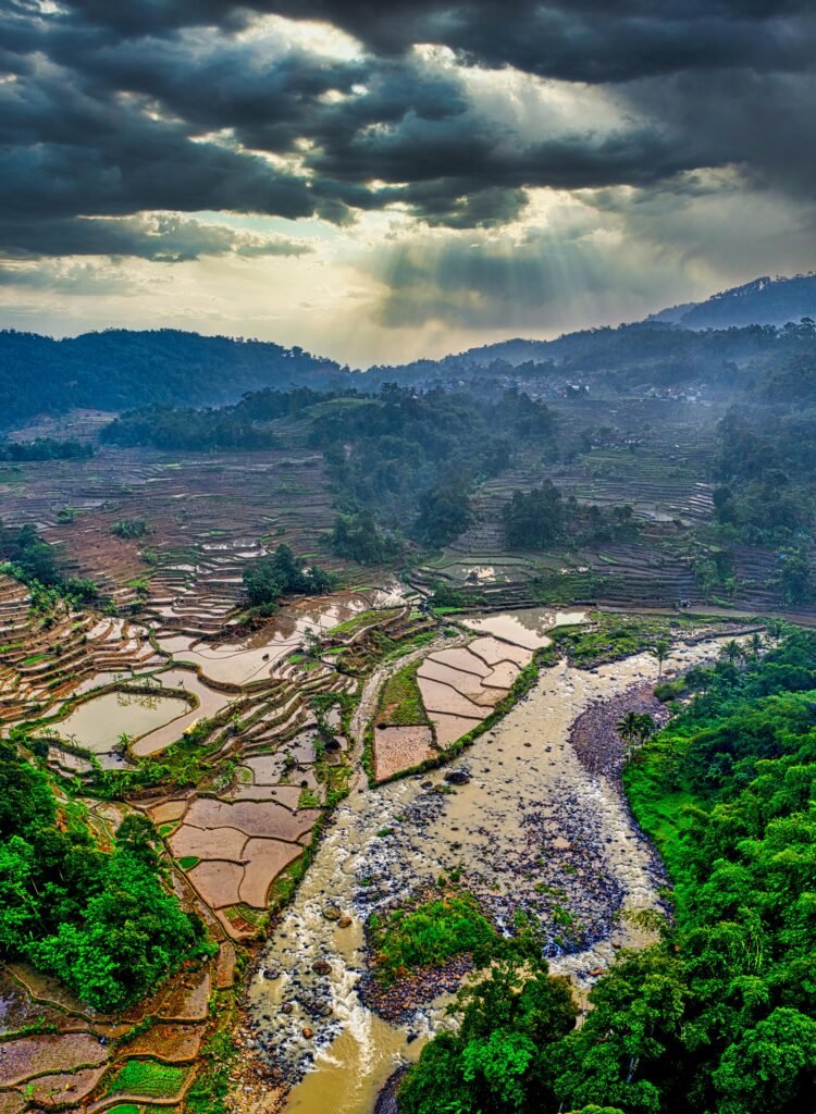 Drone shot capturing vibrant terraced rice fields and river in West Java, Indonesia under dramatic skies.